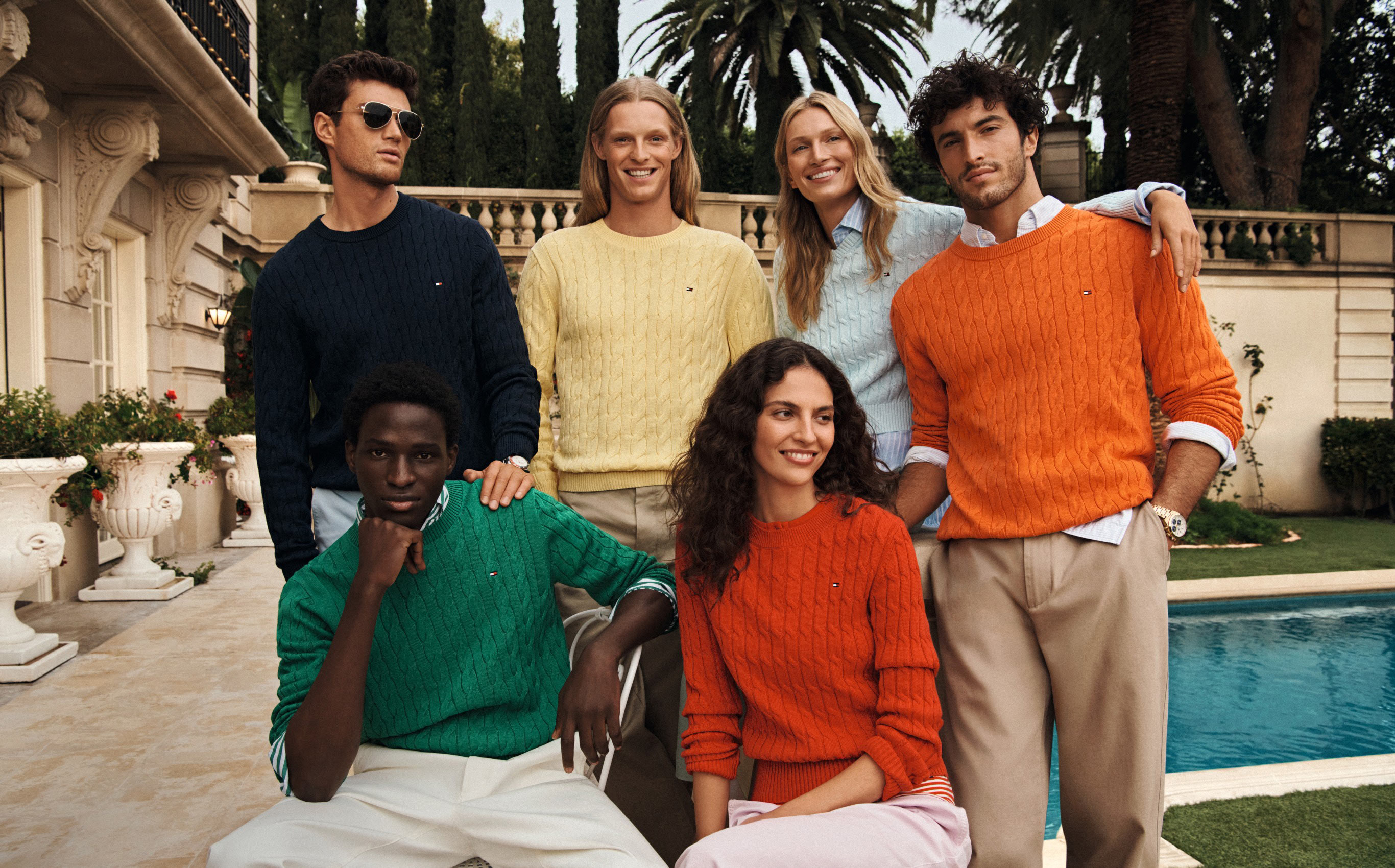 Group of models posing by a pool in colorful Tommy Hilfiger sweaters and casual separates