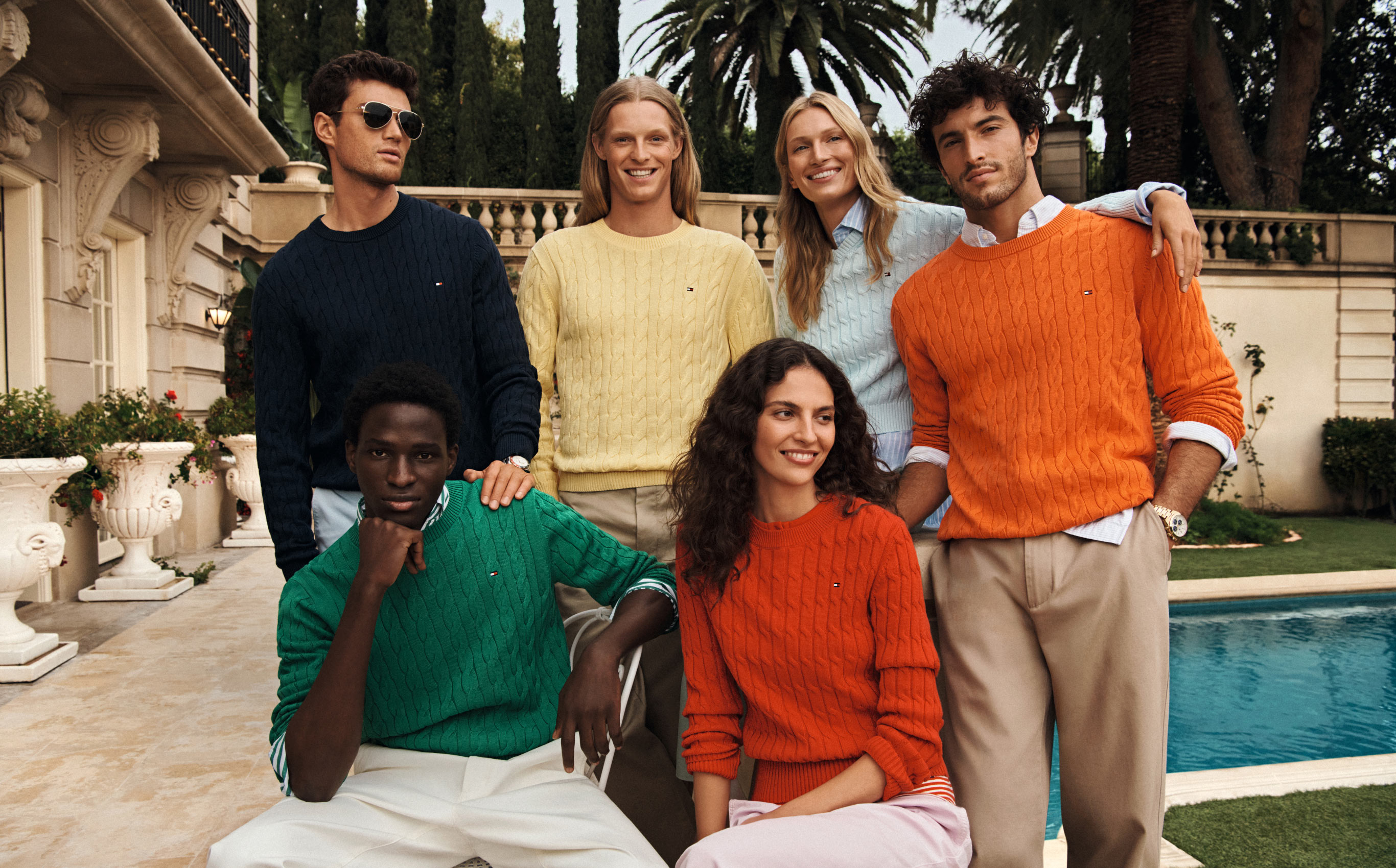 Group of models posing by a pool in colorful Tommy Hilfiger sweaters and casual separates
