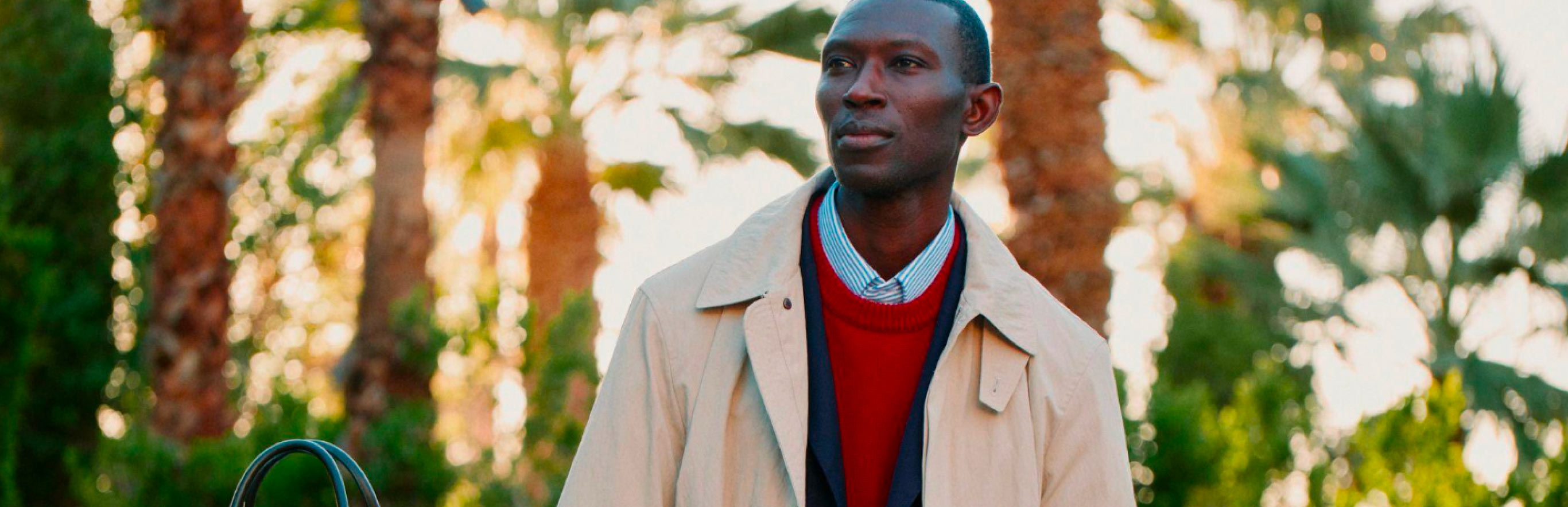 Male model outdoors wearing a beige Tommy Hilfiger trench coat over a striped sweater and jeans, holding a small bag near a parked car.