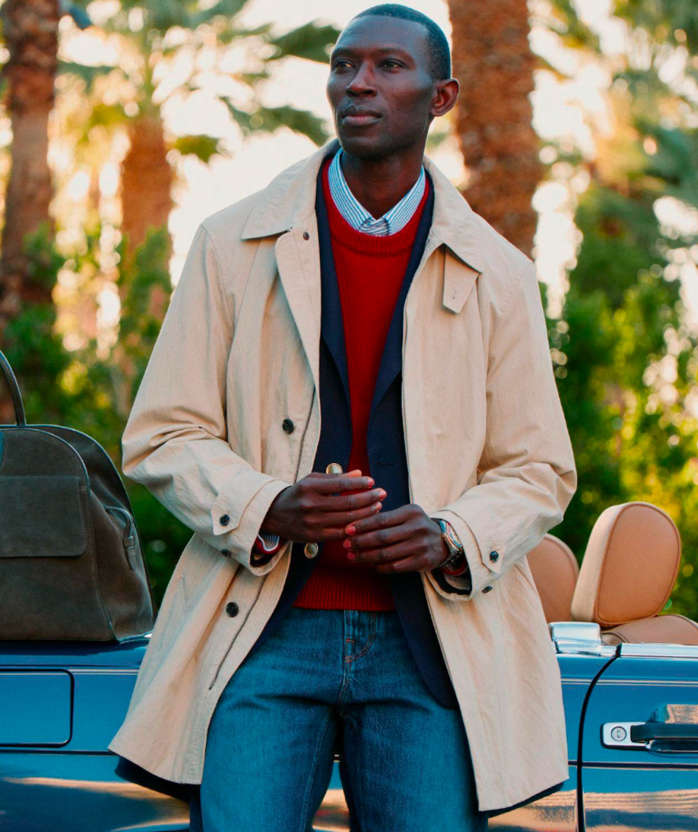 Male model outdoors wearing a beige Tommy Hilfiger trench coat over a striped sweater and jeans, holding a small bag near a parked car.
