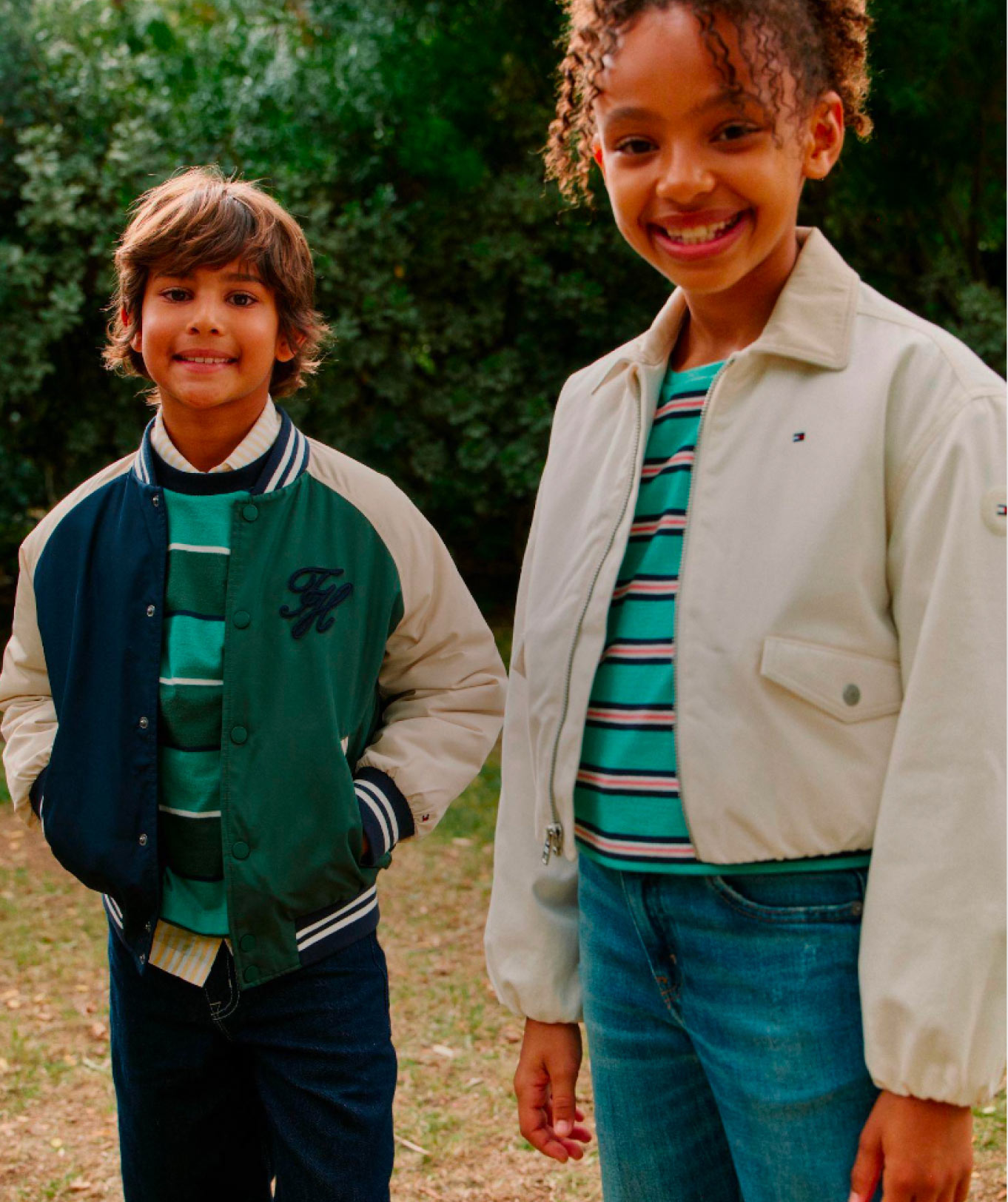 Two kids standing outdoors in casual Tommy Hilfiger outfits, with one in a green top and jacket and the other in a light jacket over a striped shirt.