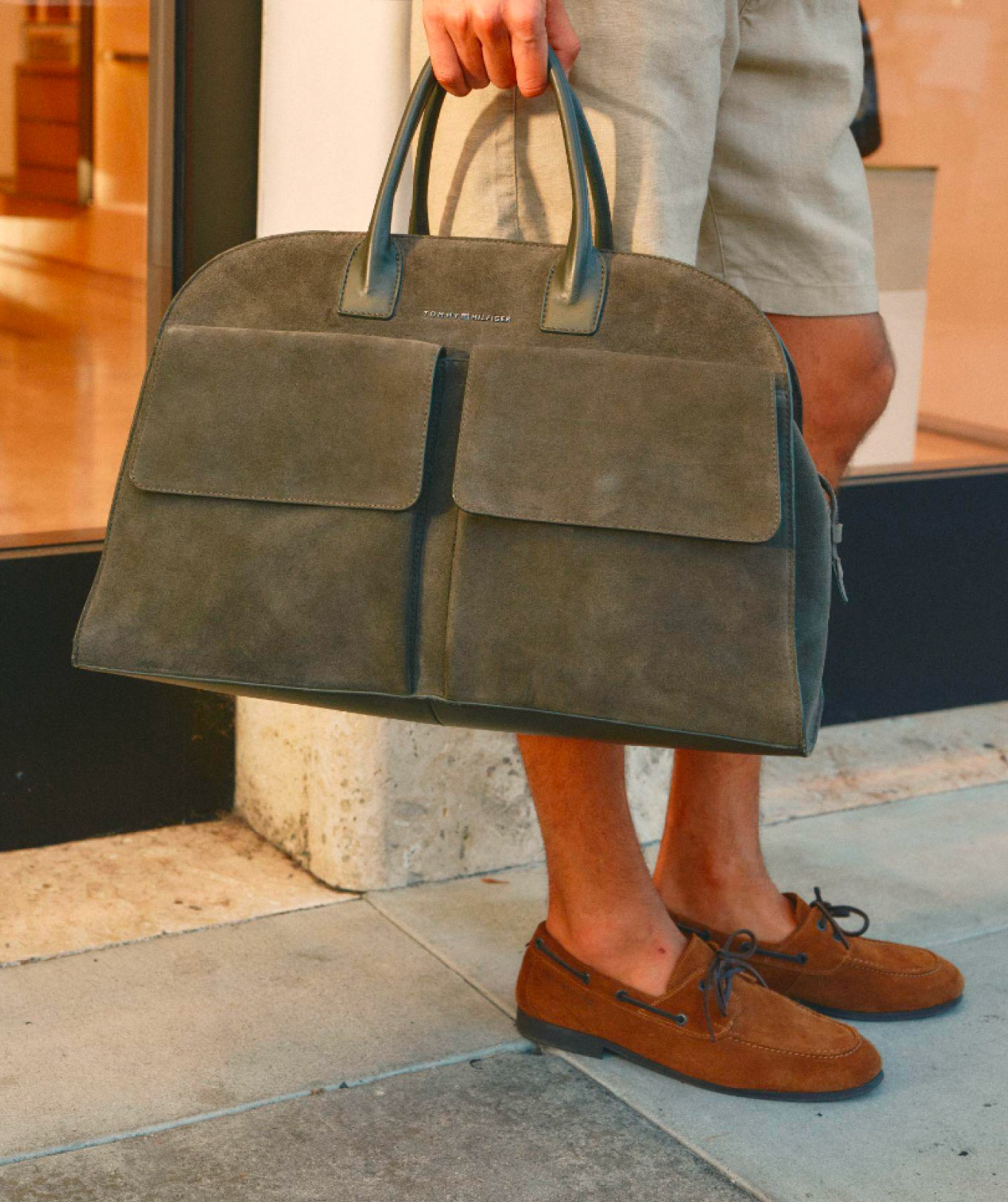 Close-up of a person holding a dark Tommy Hilfiger tote bag with 'Accessories' text overlay, standing on stone steps in loafers.