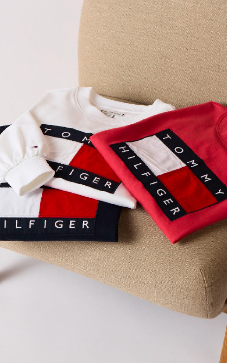 Model seated in a studio wearing a light blue Tommy Hilfiger hoodie and matching shorts with a red T-shirt peeking at the neckline.