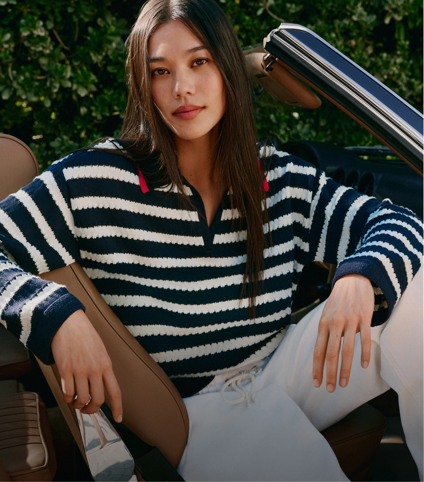 Model lounging outdoors in a navy-and-white striped Tommy Hilfiger sweater with white pants, posing beside an open-top car.