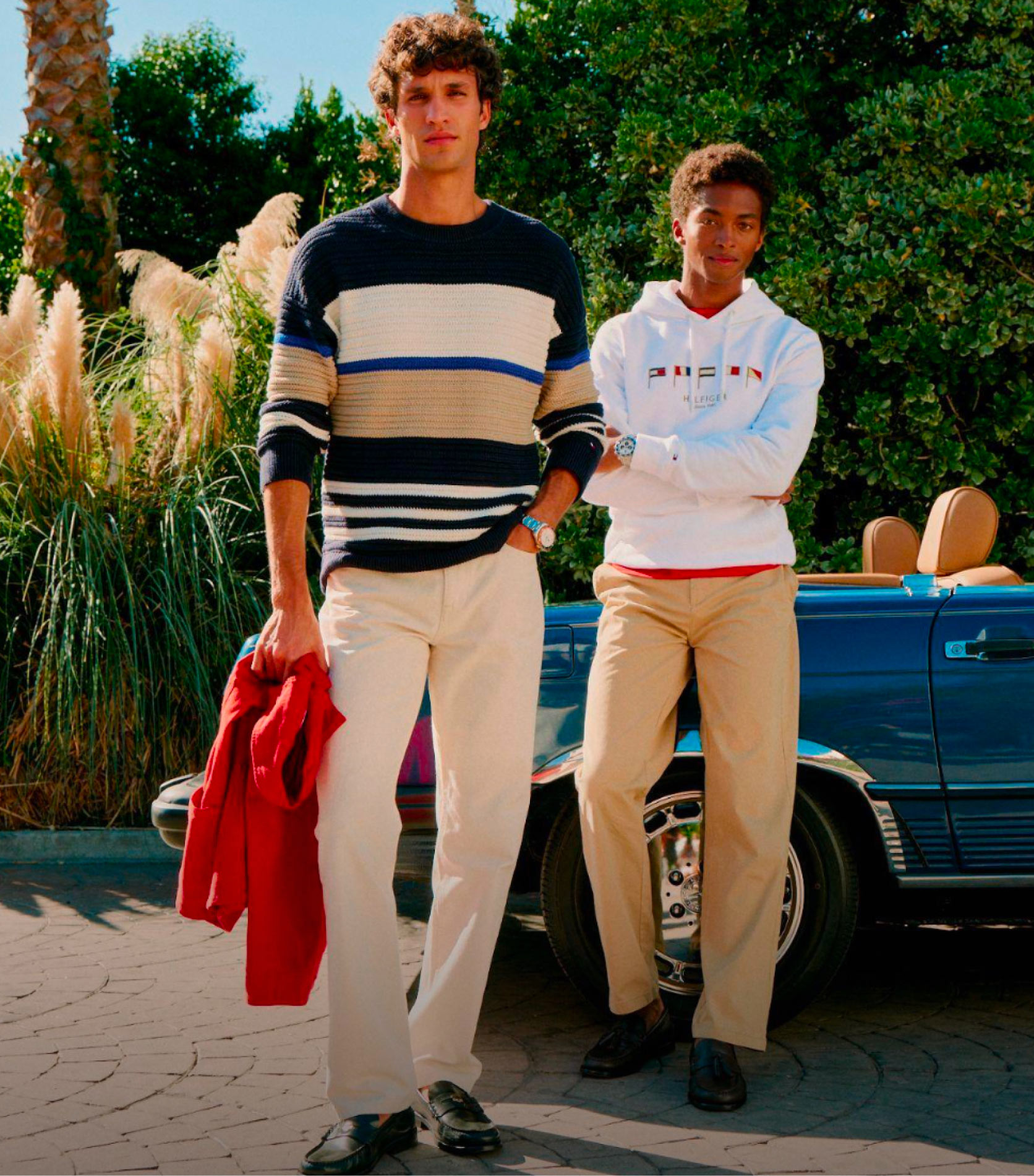 Two models standing outdoors by a convertible wearing Tommy Hilfiger chinos with casual tops, including a navy striped sweater and a white hoodie, with a red jacket carried at the side.
