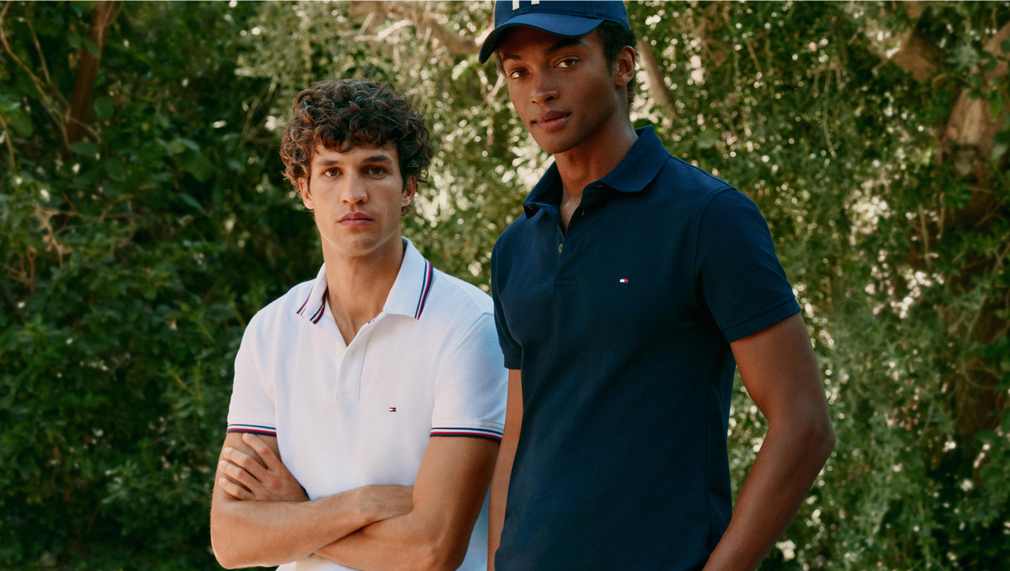 Two models posing in front of greenery wearing Tommy Hilfiger polo shirts, one in white and one in navy with a matching cap.