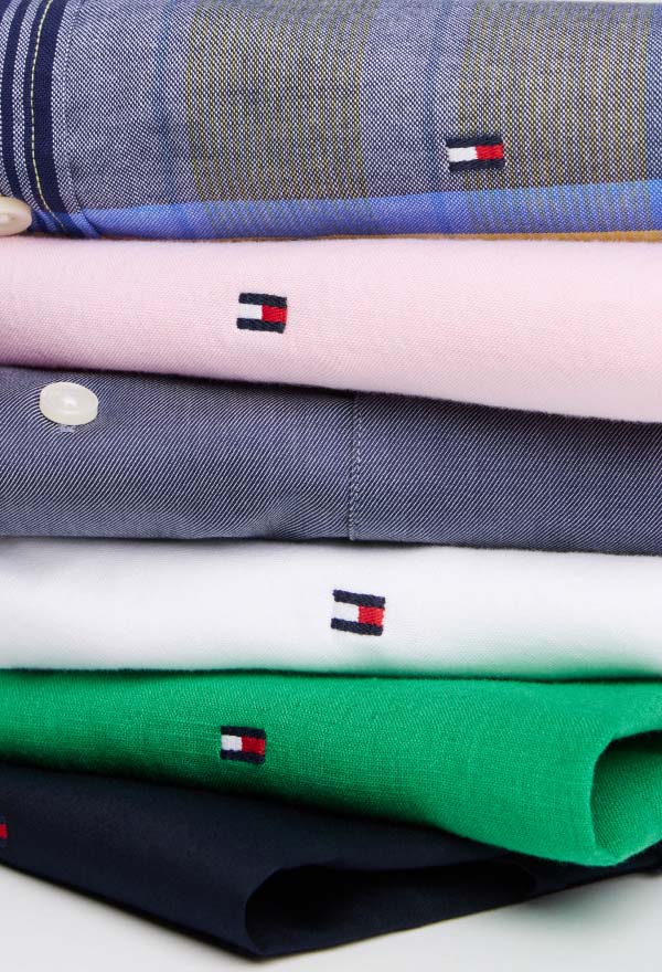 Stack of folded Tommy Hilfiger button-down shirts in various colors and patterns, including blue, white, and plaid, topped with a light blue shirt featuring a collar pin.