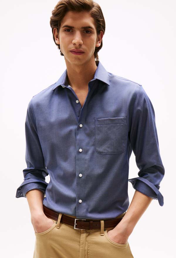 Male model wearing a blue Tommy Hilfiger button-down shirt with sleeves rolled, styled with tan chinos on a clean studio background.