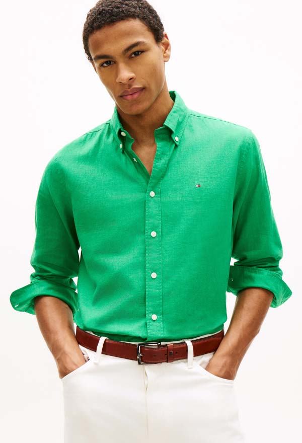 Model wearing a bright green Tommy Hilfiger long-sleeve button-down shirt with rolled sleeves and a small chest logo, styled with a brown belt and light pants in a studio setting.