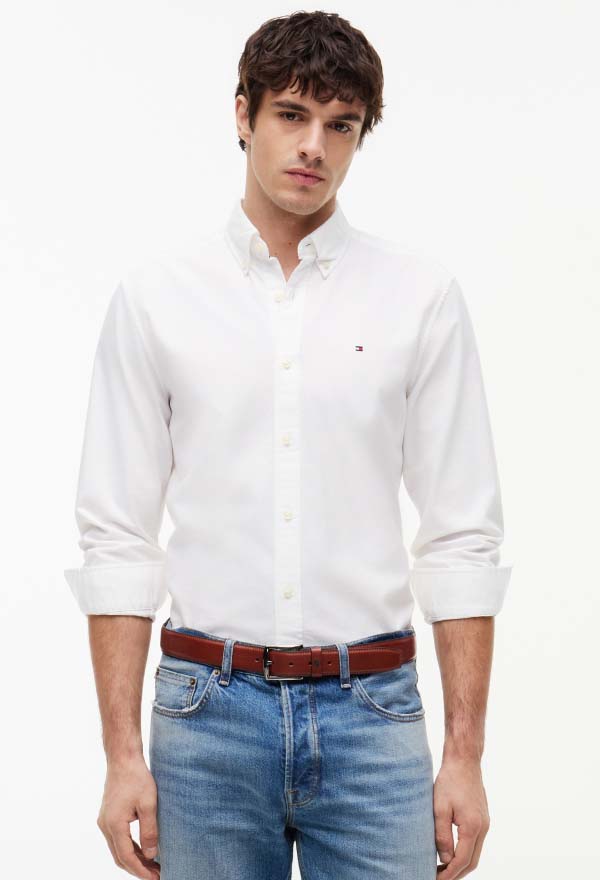 Male model wearing a white Tommy Hilfiger long-sleeve button-down shirt with a small chest flag logo, styled with a brown leather belt and light blue jeans against a plain studio background.