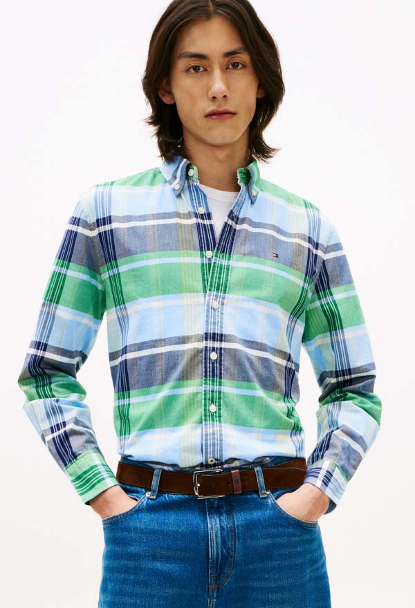 Model wearing a green and blue plaid Tommy Hilfiger button-down shirt with a small chest logo, styled with blue jeans and a brown belt in a studio setting.