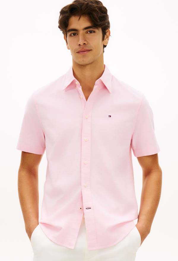 Model wearing a light pink Tommy Hilfiger short-sleeve button-down shirt with a small chest flag logo, paired with white pants in a studio setting.