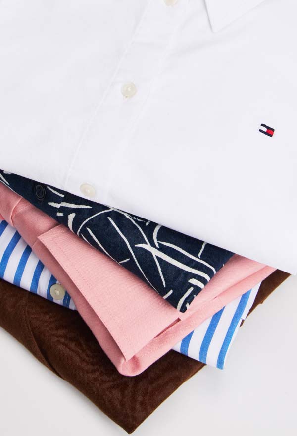 Stack of folded Tommy Hilfiger women's shirts in various styles and fabrics, including white, navy blue, pink, blue-striped, and deep red, showcasing collars and button details.