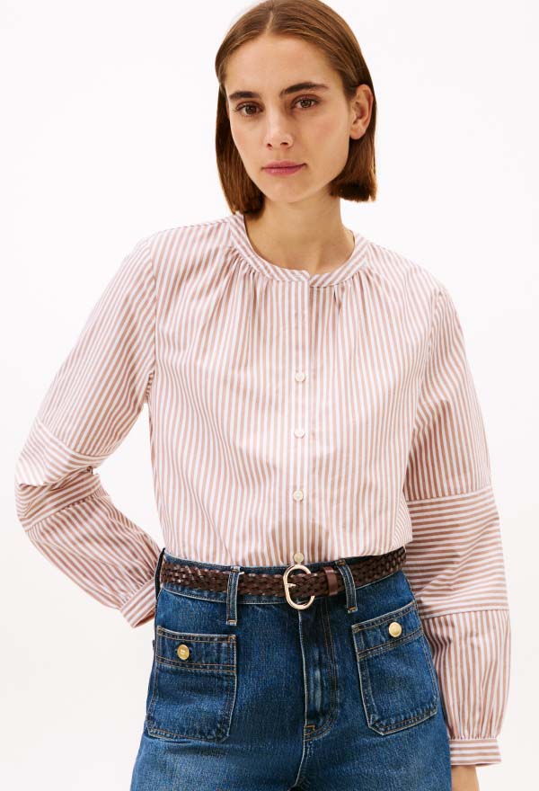 Model wearing a pink striped Tommy Hilfiger blouse with gathered neckline and long sleeves, styled with blue jeans and a black belt in a studio setting.