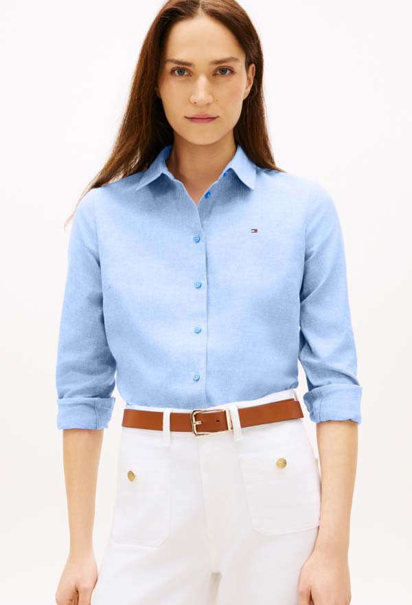 Model wearing a light blue Tommy Hilfiger button-down shirt with a small chest logo, styled with white pants and a tan belt in a studio setting.
