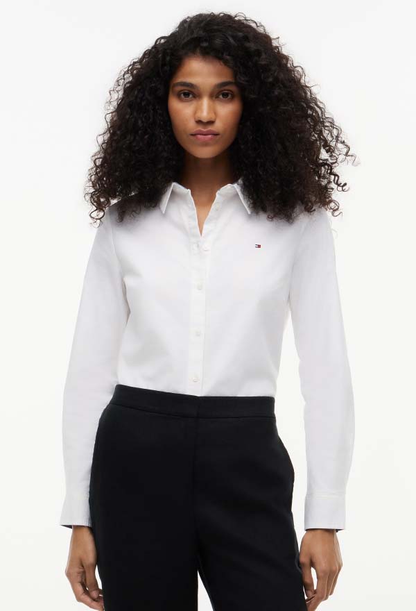 Model wearing a white Tommy Hilfiger button-down shirt with a small chest flag logo, styled with black trousers on a clean studio background.