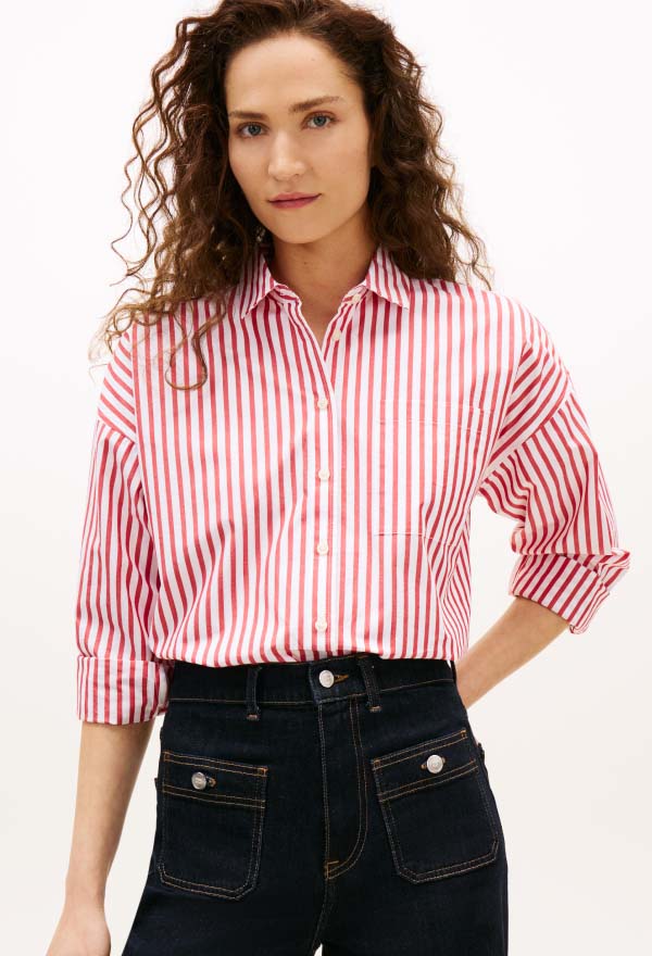 Model wearing a red-and-white striped Tommy Hilfiger button-down shirt with sleeves rolled, styled with dark denim jeans in a studio setting.