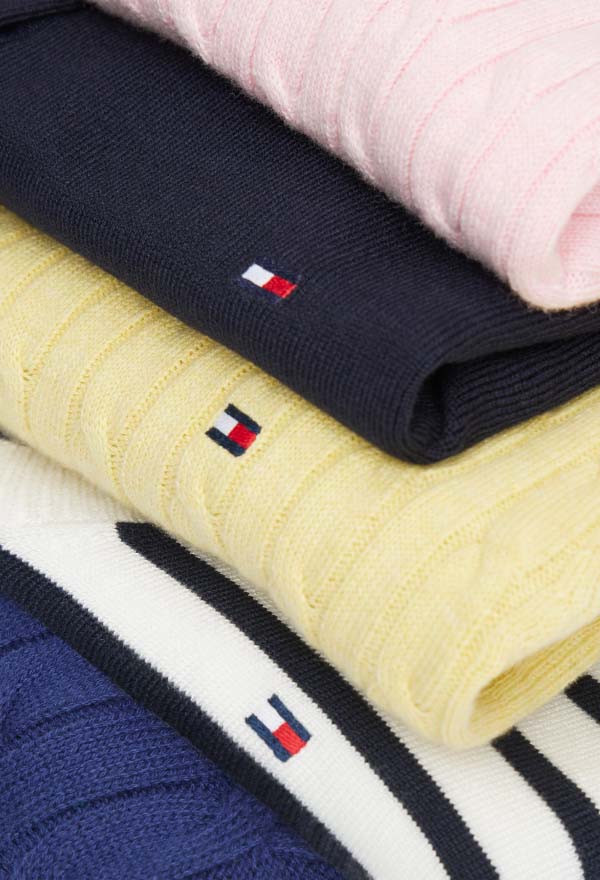 Close-up of folded Tommy Hilfiger knit sweaters in pastel pink, navy, and light yellow, showing small embroidered flag logos on a neutral surface.