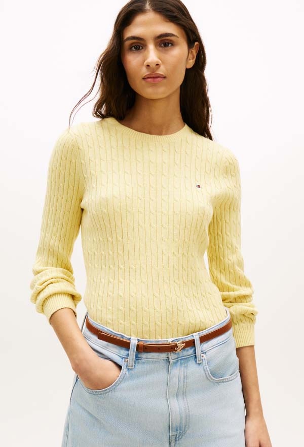 Model wearing a light yellow Tommy Hilfiger crewneck sweater with a small chest flag logo, styled with jeans and a brown belt against a clean studio background.