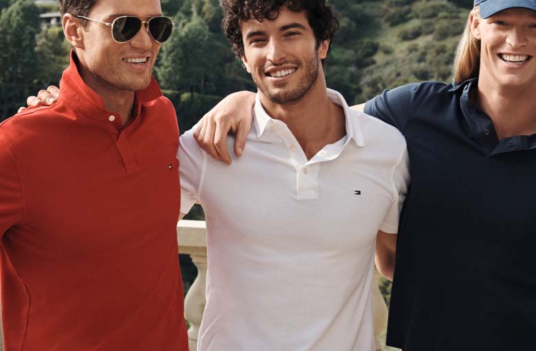 Group of three models standing outdoors with arms around each other wearing Tommy Hilfiger polo shirts in red, white, and navy, styled with sunglasses and a baseball cap in a sunny setting.