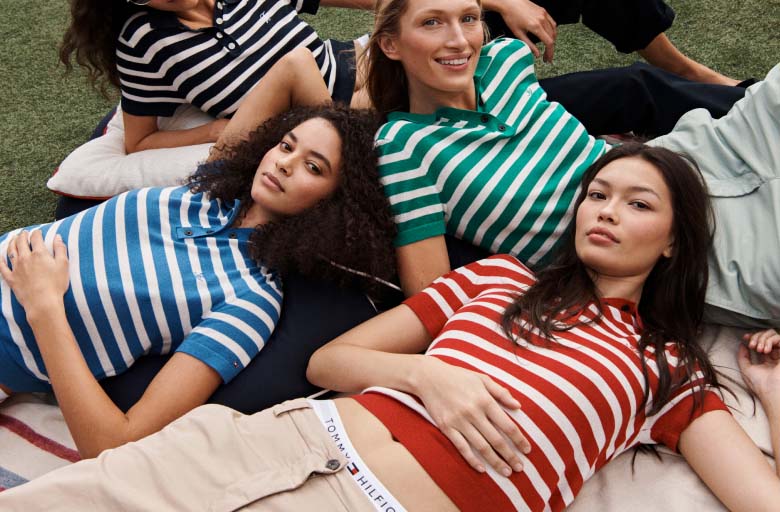 Group of models lying on grass outdoors wearing striped Tommy Hilfiger sweaters in red, green, blue, and black, styled with casual pants and relaxed poses in a sunny setting.