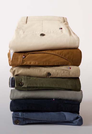 Folded stack of neutral-toned Tommy Hilfiger chino pants arranged vertically, showing a range of classic colors against a clean studio background.