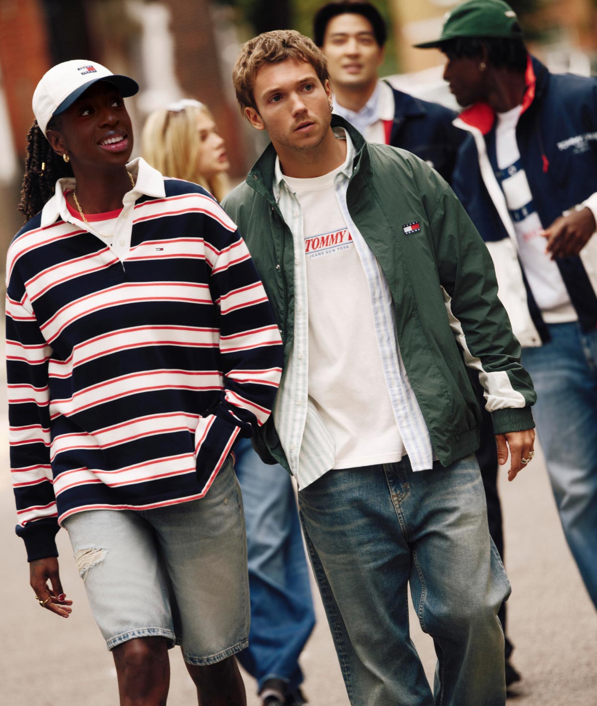 Group of models walking outdoors in casual Tommy Hilfiger looks, including a navy, red, and white striped rugby shirt with denim shorts and a green jacket layered over a white T shirt with jeans.