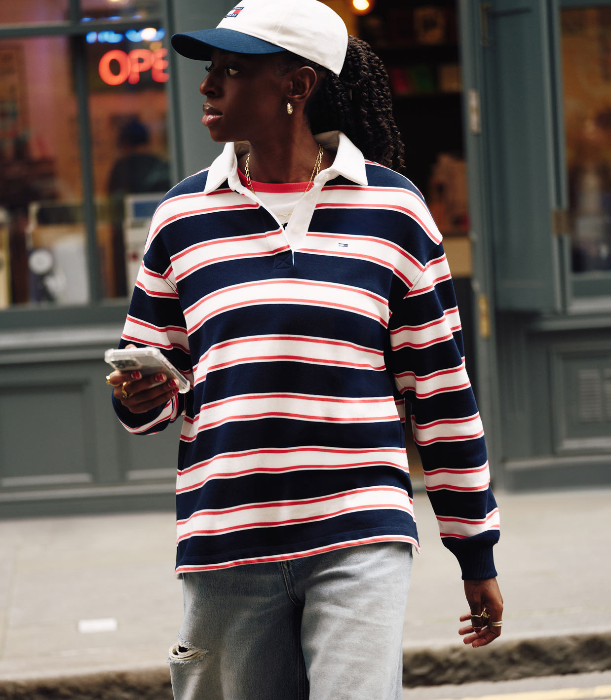 Model walking outdoors wearing a navy, red, and white striped Tommy Hilfiger rugby shirt with a white cap and light denim shorts.