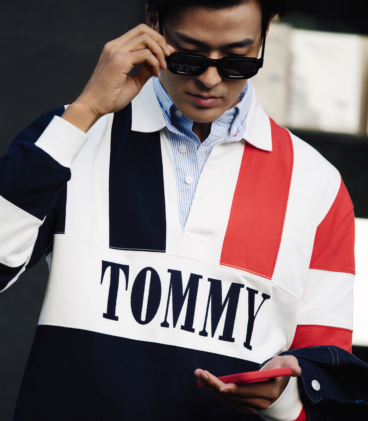 Close up of model adjusting sunglasses wearing a colorblocked Tommy Hilfiger pullover with a large TOMMY logo over a light blue button down shirt.