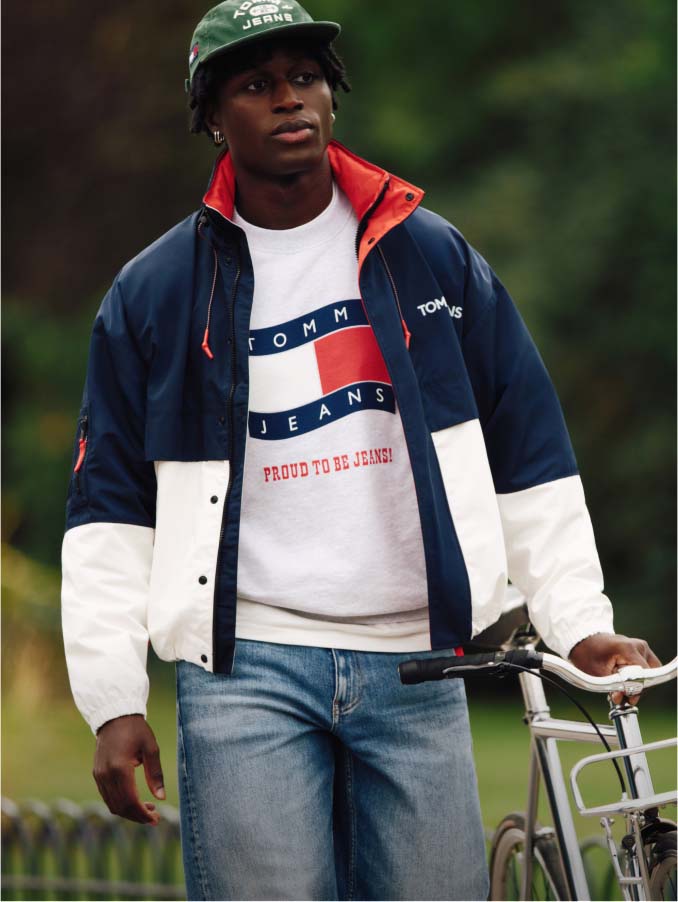 Model walking next to bike wearing Tommy Jeans colorblock jacket and sweatshirt.