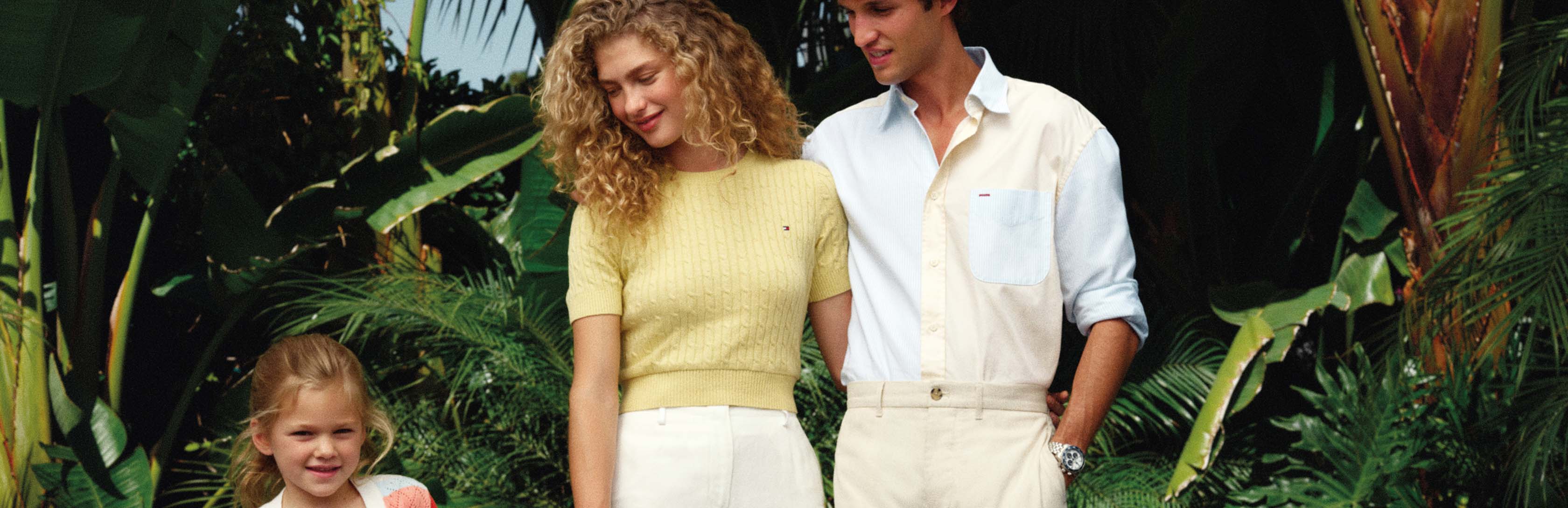 Family walking through a leafy outdoor patio setting in coordinated Tommy Hilfiger looks, including cream trousers, a pale yellow top, and a colorful striped shirt on the child.