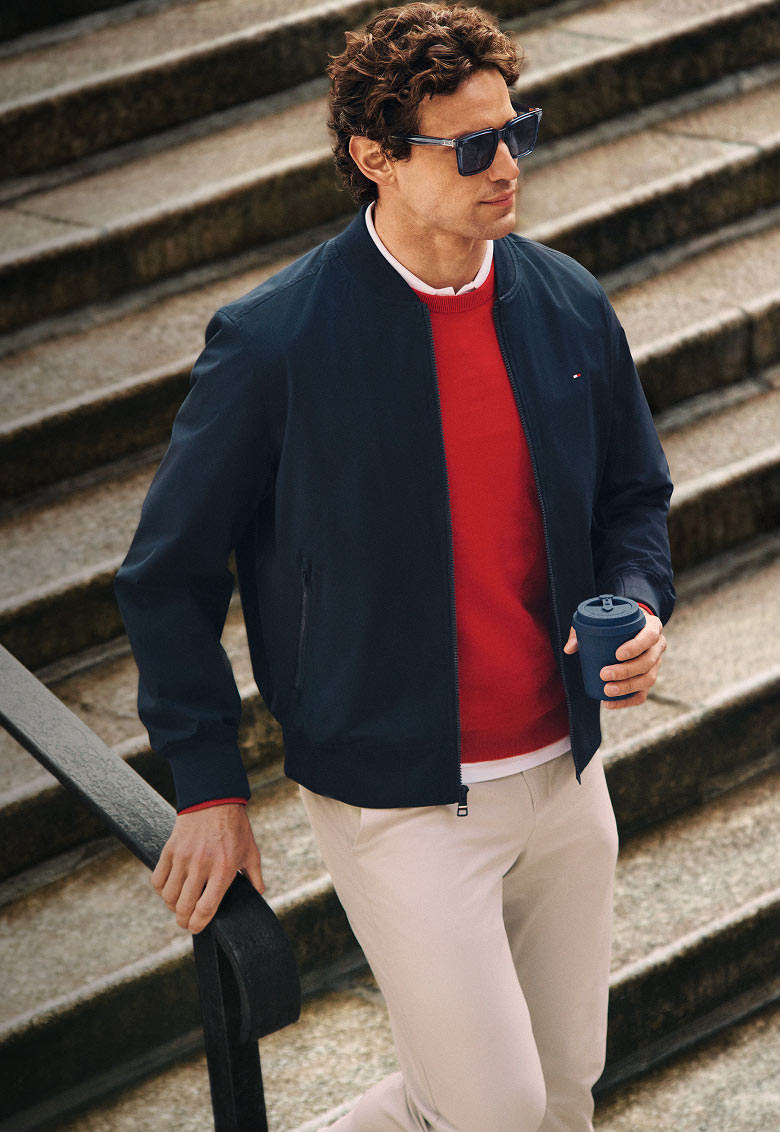 Model outdoors in casual Tommy Hilfiger looks, including a navy jacket layered over a red sweater with beige trousers.