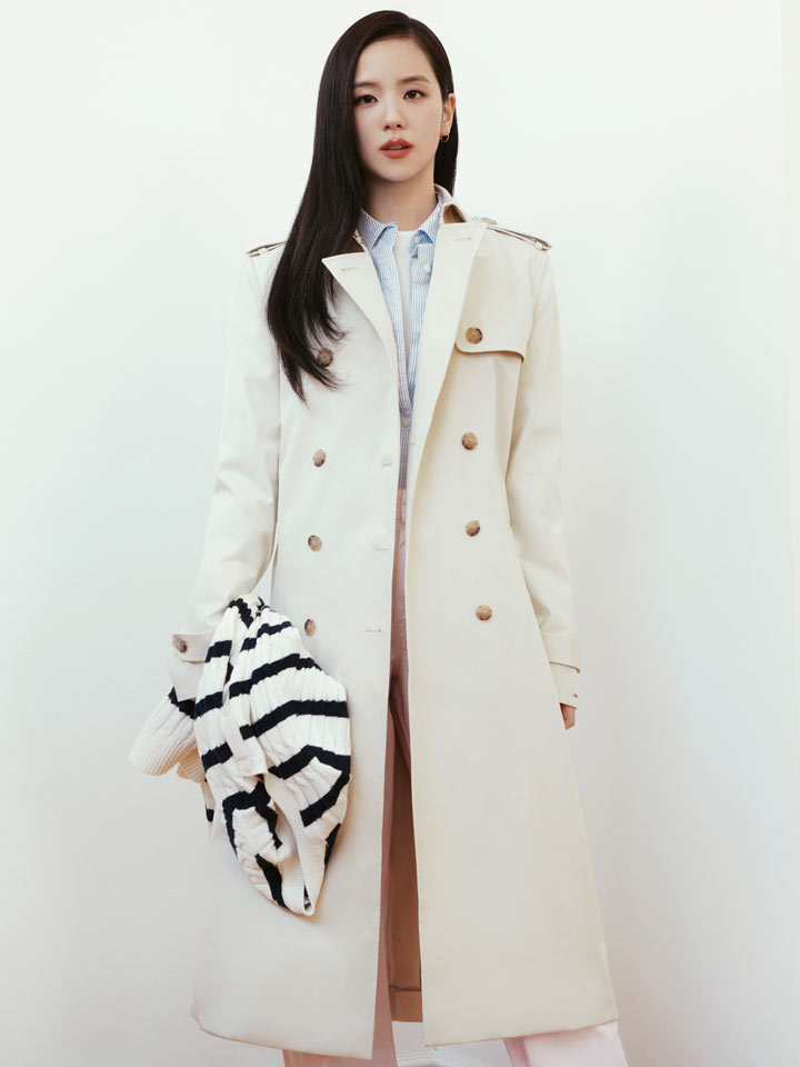 Full body shot of Jisoo wearing a long cream Tommy Hilfiger coat over a black and white patterned top, paired with wide leg white trousers and white shoes, standing indoors against a light wall.