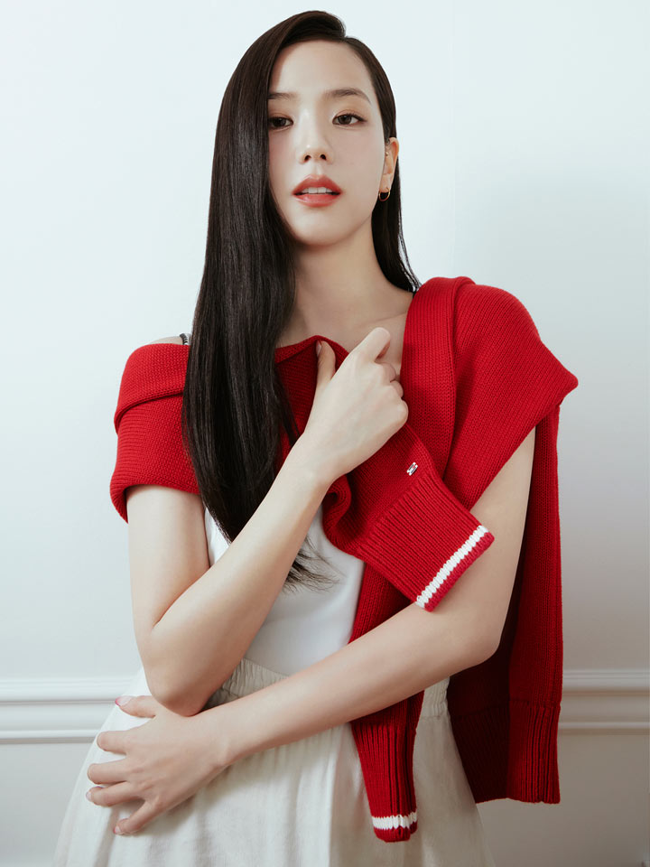 Close up portrait of Jisoo wearing a red Tommy Hilfiger short sleeve top, seated with knees drawn up against a plain studio backdrop.