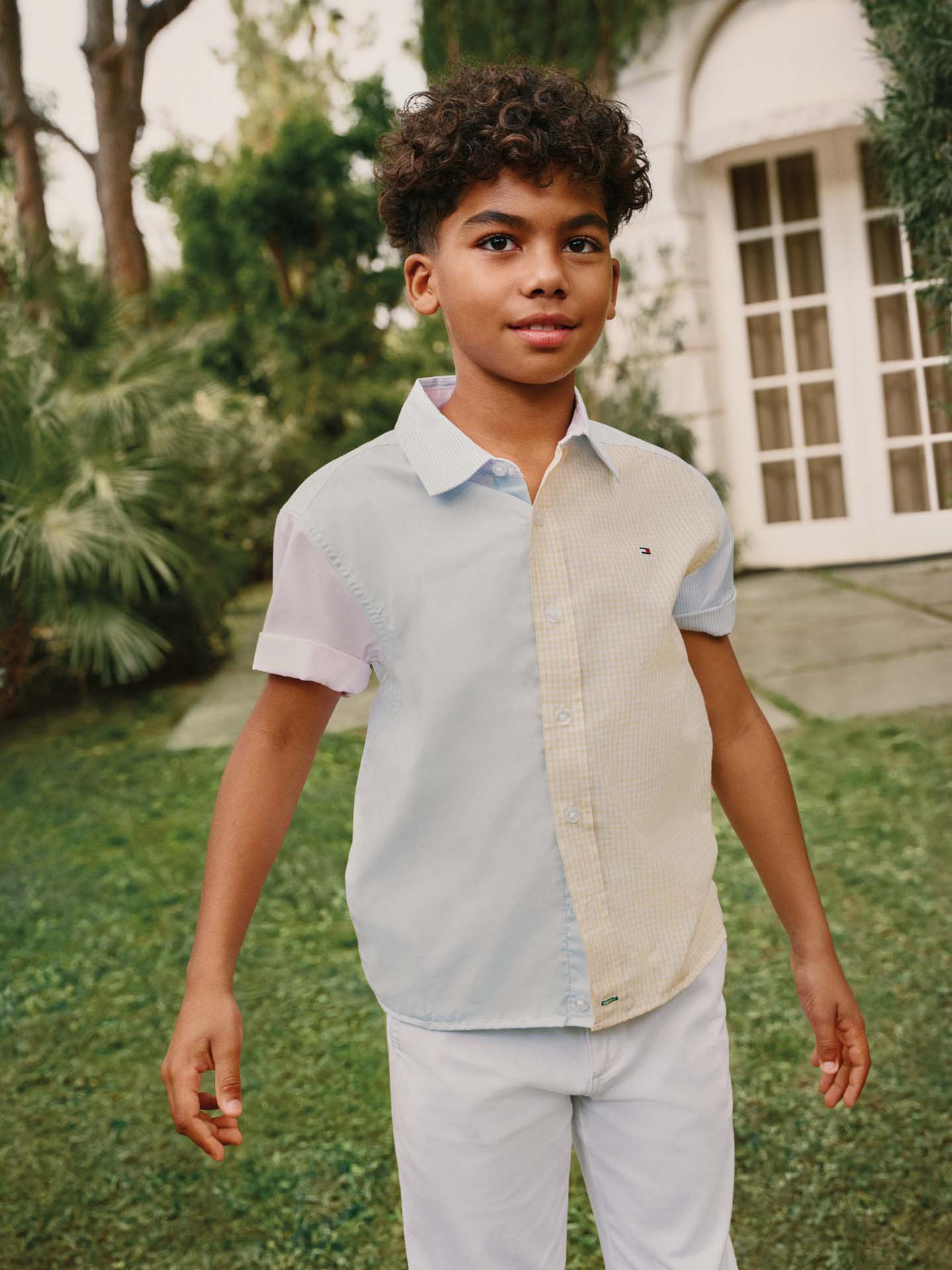 Child walking on grass outdoors wearing a color-block Tommy Hilfiger short-sleeve polo shirt in light blue, beige, and white, paired with light shorts.
