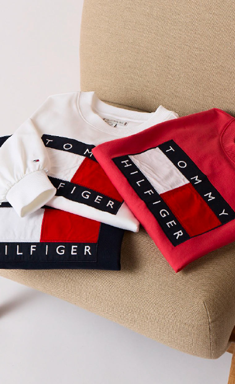 Folded Tommy Hilfiger pieces arranged on a beige surface, including a red garment and a white collared shirt with branded tape and logo details.