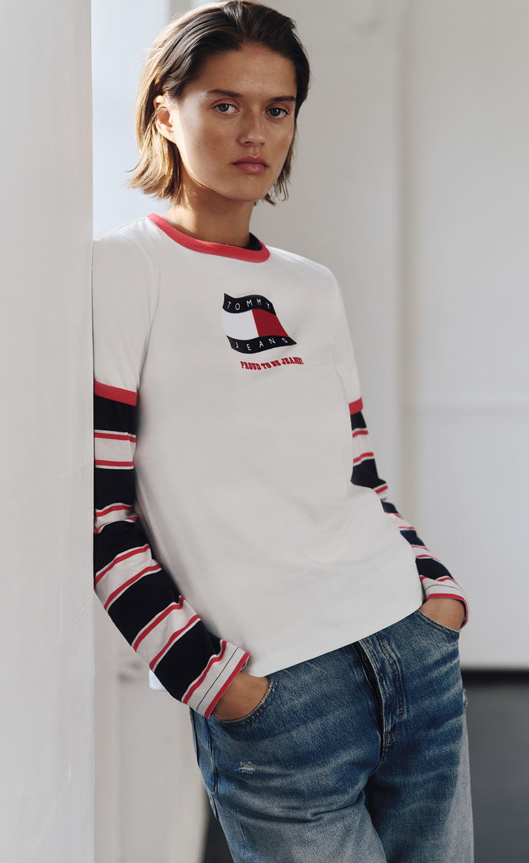 Model leaning indoors in a white Tommy Hilfiger sweater with a large flag logo and striped sleeves, paired with blue jeans.