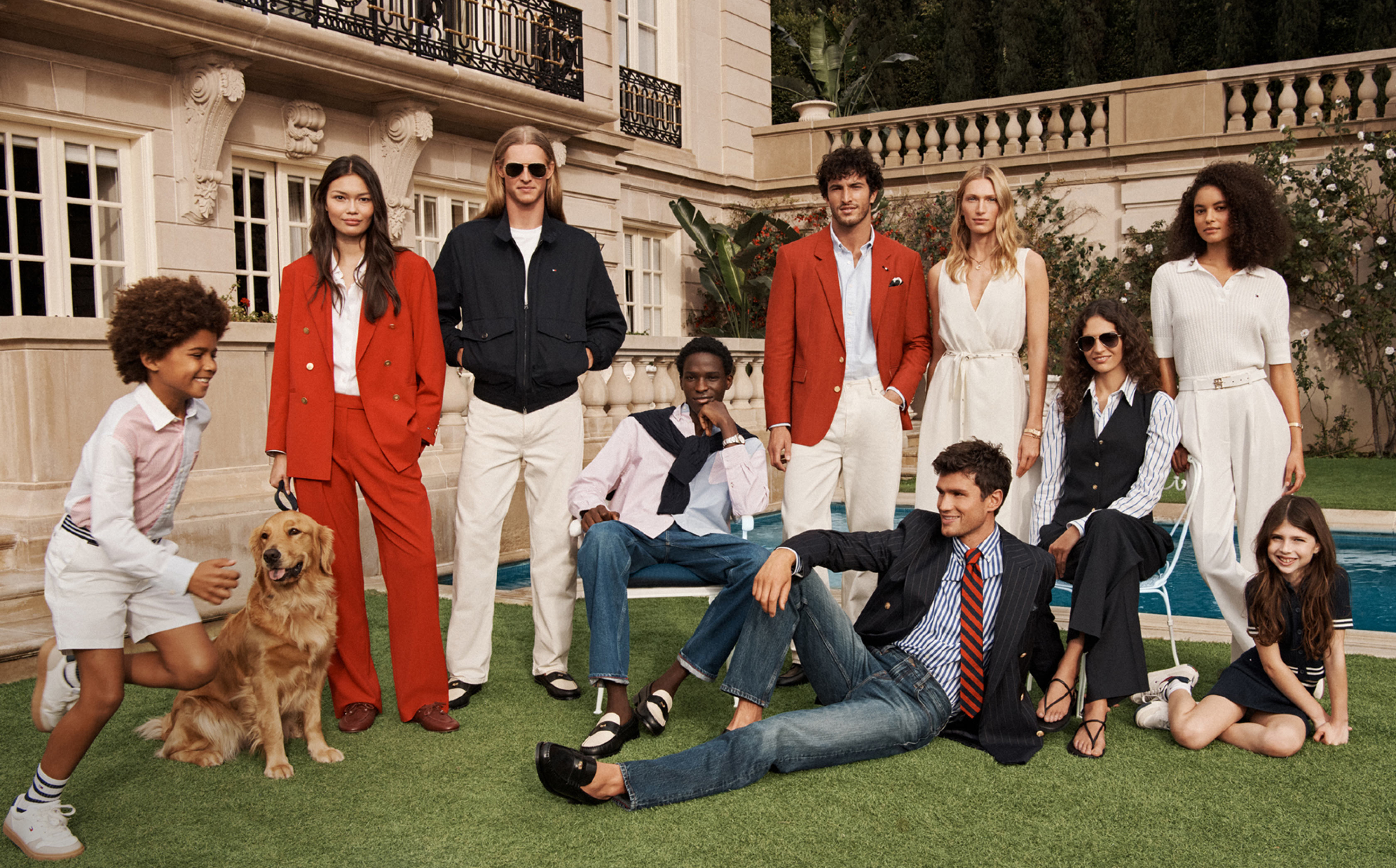 Group of models lounging on a lawn outside a building wearing mixed Tommy Hilfiger outfits, including a bright red dress, light suits, and casual separates.