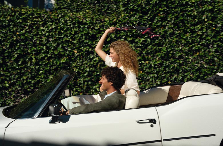 Woman riding in a convertible raising her arm, with curly hair and a light Tommy Hilfiger top visible above the car door.