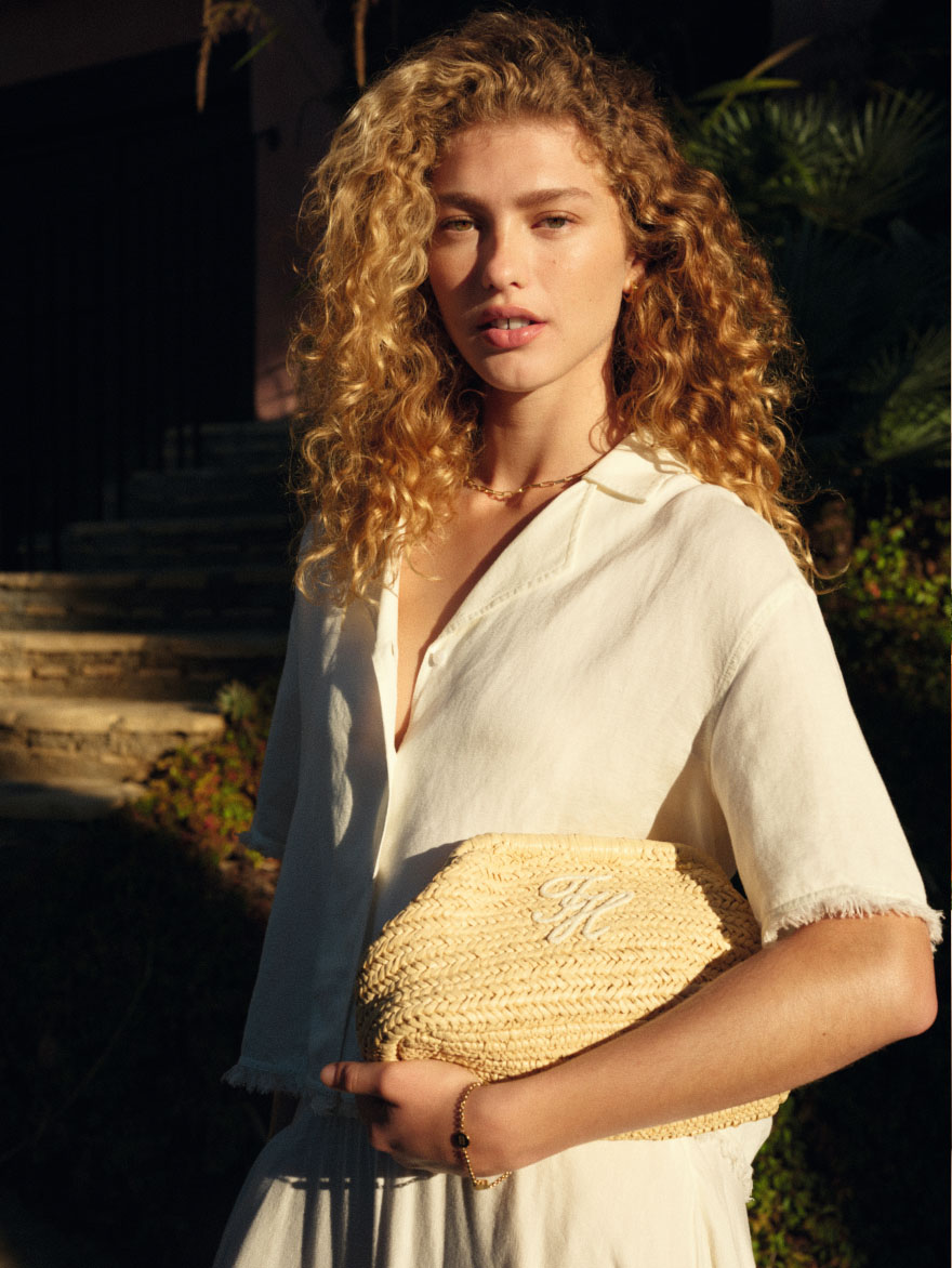 Model posed outdoors in warm light wearing a cream Tommy Hilfiger blouse, holding a woven straw-style clutch.