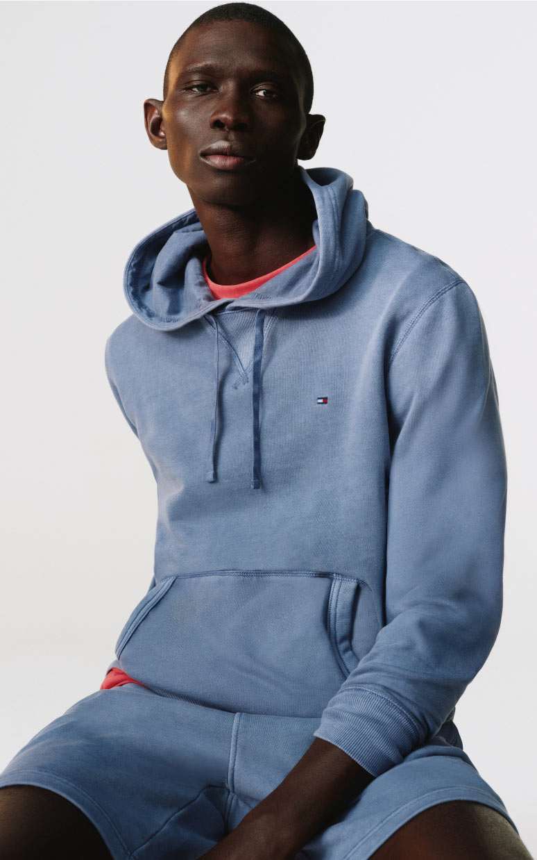 Model seated in a studio wearing a light blue Tommy Hilfiger hoodie and matching shorts with a red T-shirt peeking at the neckline.