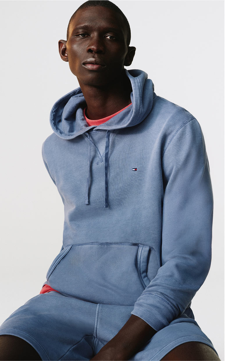 Model seated in a studio wearing a light blue Tommy Hilfiger hoodie and matching shorts with a red T-shirt peeking at the neckline.