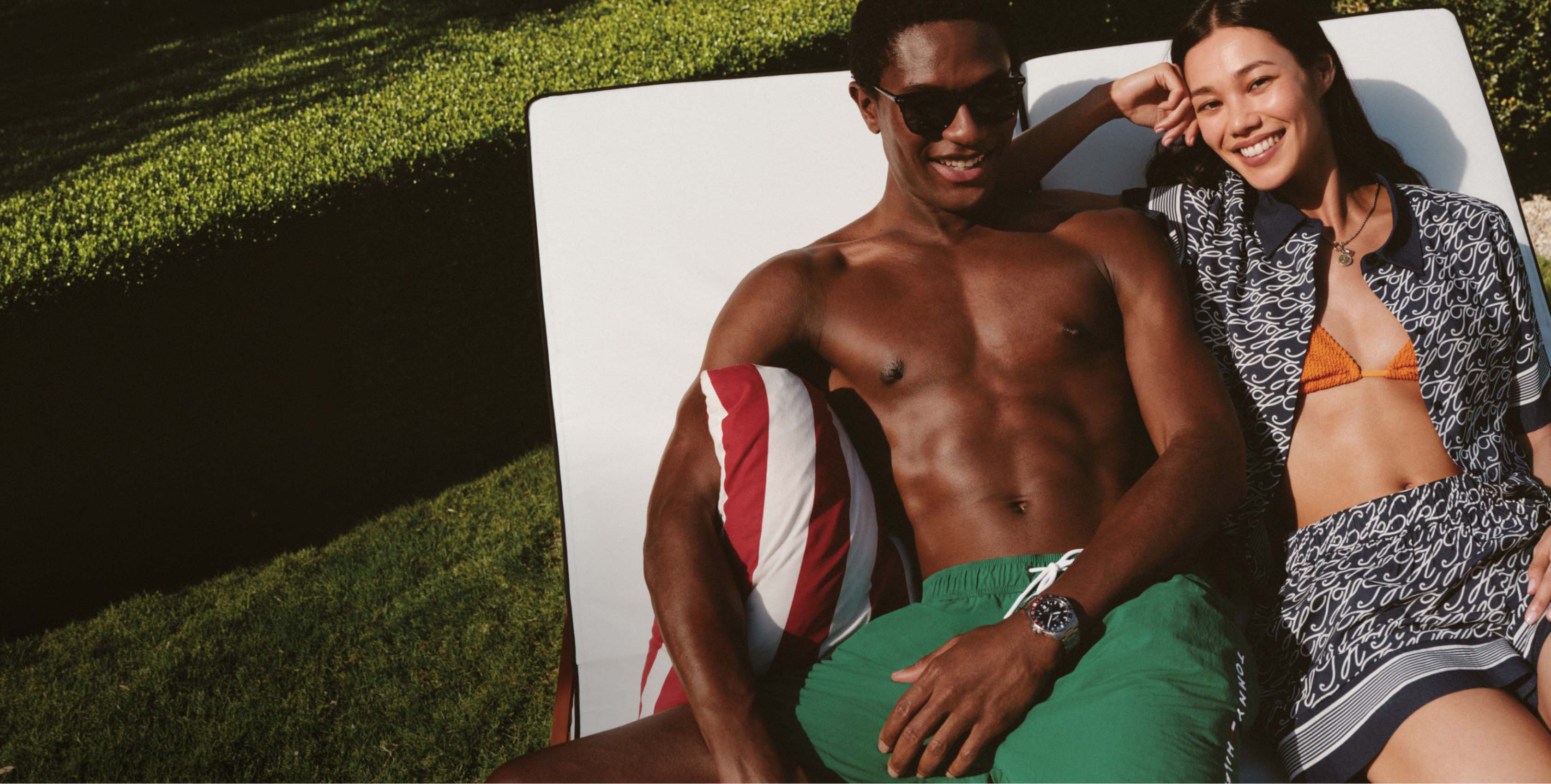 Man and woman lounging poolside on chairs, wearing Tommy Hilfiger swimwear and sunglasses in a sunny outdoor setting.