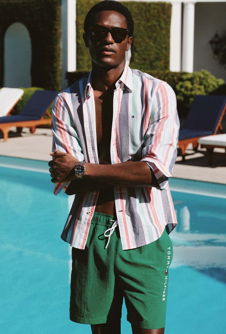 Man standing by a pool wearing a Tommy Hilfiger striped shirt and green swim shorts.