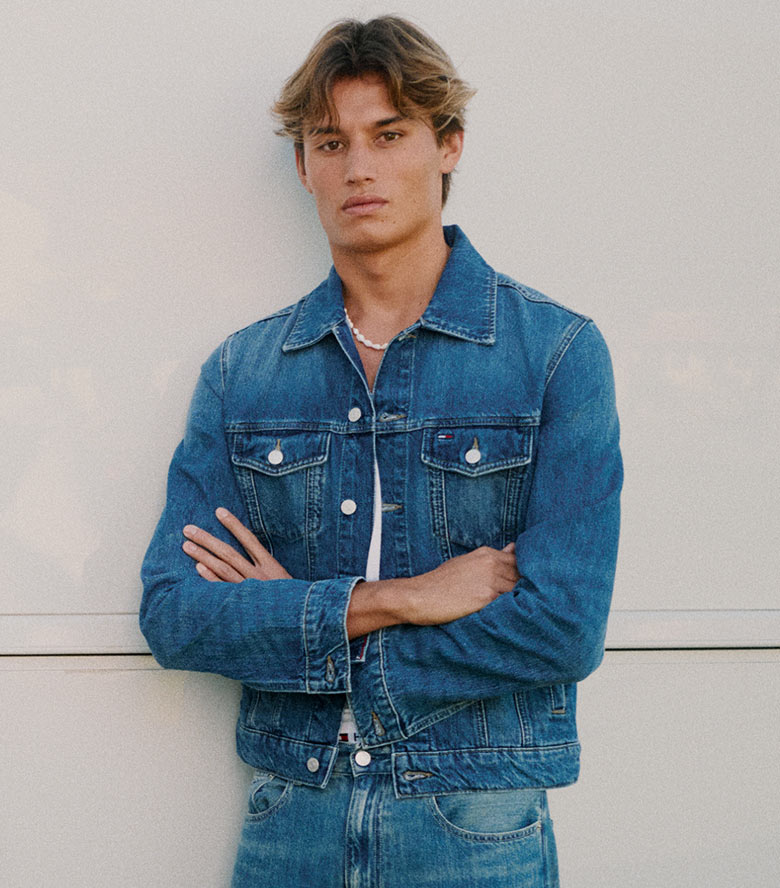 Close up of model adjusting sunglasses wearing a colorblocked Tommy Hilfiger pullover with a large TOMMY logo over a light blue button down shirt.