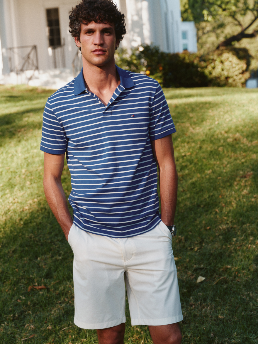 A man wearing a blue Tommy Hilfiger striped polo shirt and white shorts standing on a grassy lawn.