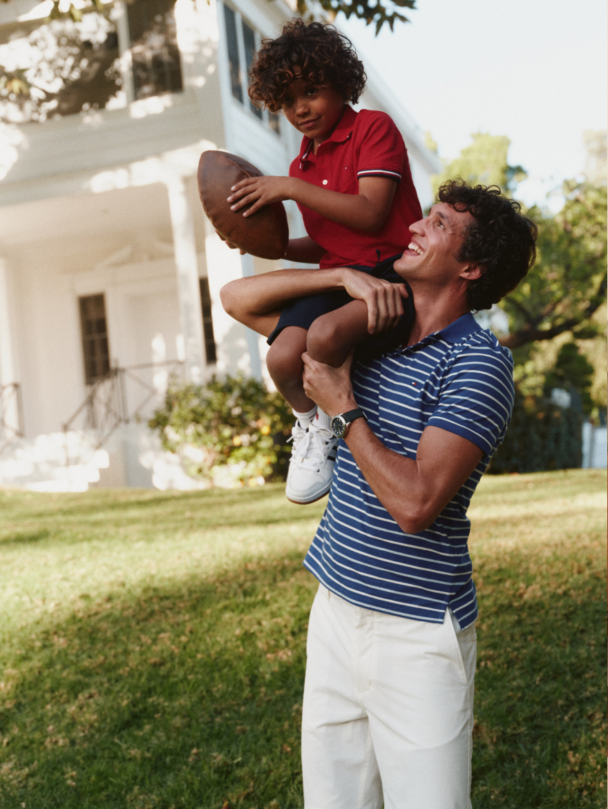 A man carrying a young boy on his shoulders while the boy holds a football, both outdoors on a lawn wearing Tommy Hilfiger.