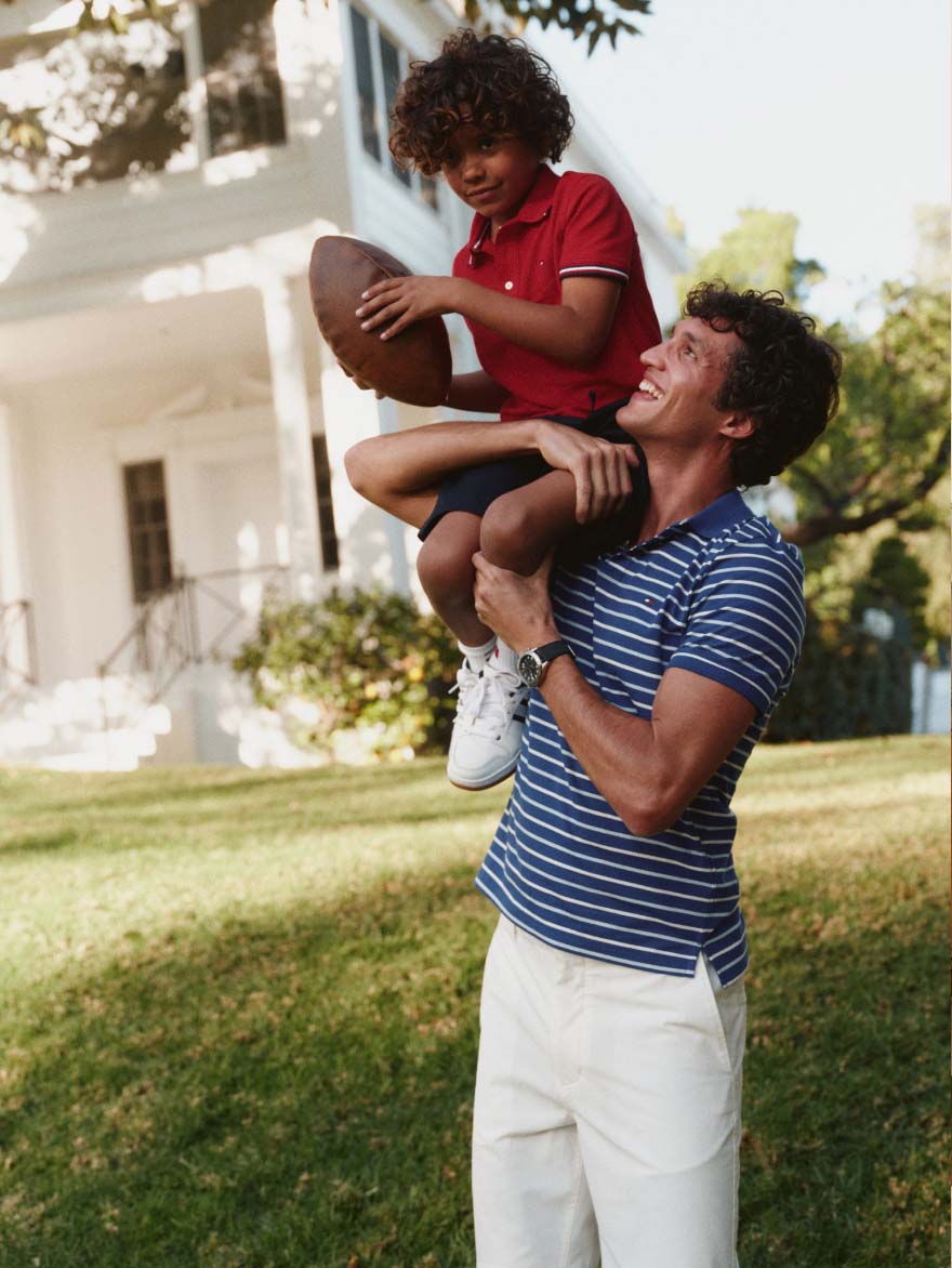 A man carrying a young boy on his shoulders while the boy holds a football, both outdoors on a lawn wearing Tommy Hilfiger.