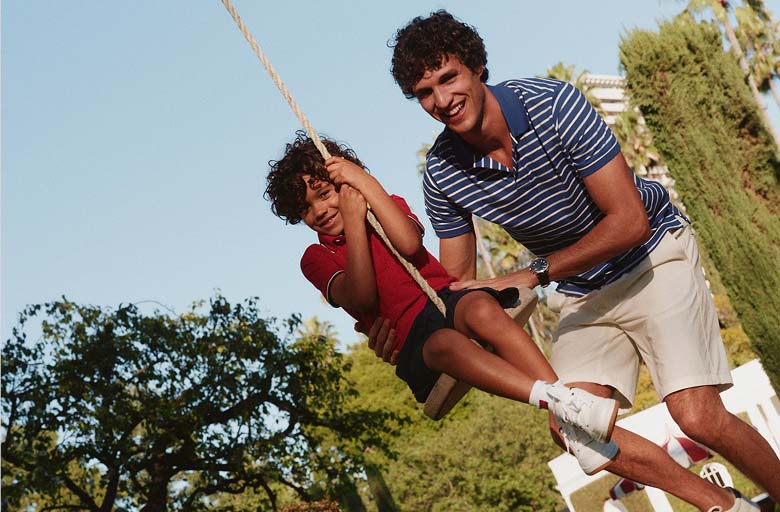 A man and a young boy smiling while holding onto a rope outdoors on a sunny day wearing Tommy Hilfiger outfits.