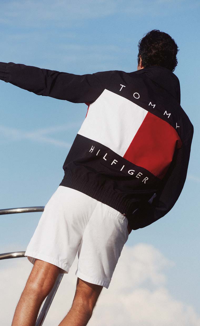 Man standing on a boat with arms outstretched, wearing a Tommy Hilfiger navy jacket with a large red, white, and blue Tommy Hilfiger logo on the back against a blue sky.